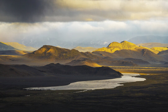 Aerial view of Landmannalaugar featuring volcanic mountains illuminated by golden hour light, a winding river, and dark dramatic clouds in Rangarthing ytra, Iceland.