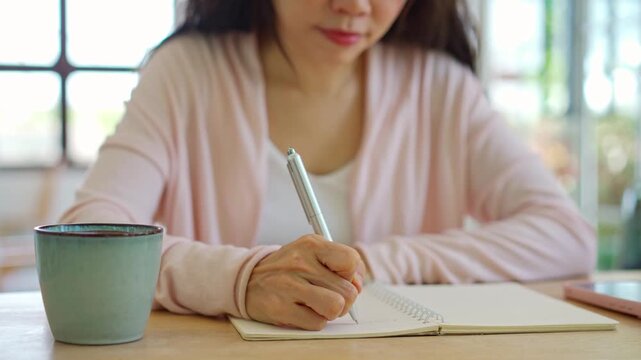 Smiling Asian woman writing down ideas in a notebook during work or study in a bright cozy room