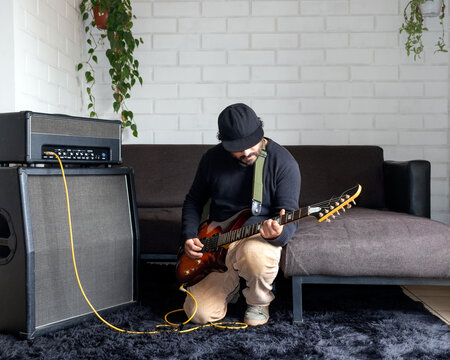 Latino musician kneeling while playing a electric guitar next to an amplifier at home