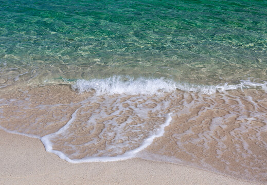 Emerald sea shoreline. Clear green water laps fine sand. Small waves leave foamy patterns on the beach.