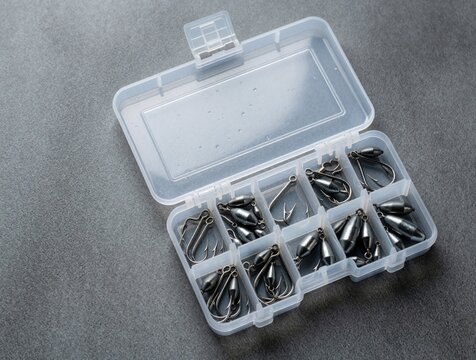 Transparent plastic box holds metal fishing hooks, lead sinkers. Organizer tray stores essential angling gear on grey surface. Open portable container displays assorted terminal tackle equipment for