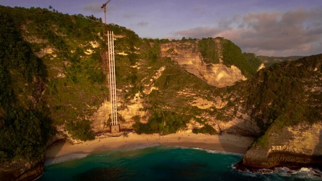 Aerial view of Kelingking Beach with a tall construction elevator structure against the cliff face at sunset in Nusa Penida, Bali, Indonesia.