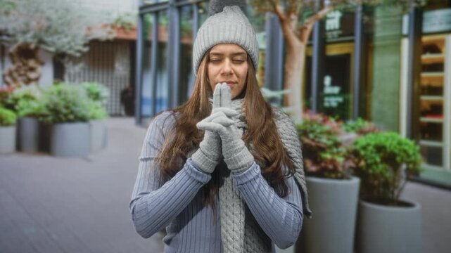 Woman in gray beanie and knit scarf clasps gloved hands near her face with subtle expression on street storefront; contemplation warmth.