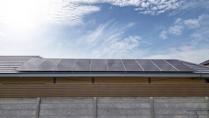 Fototapeta premium Residential rooftop solar panels installed on a house tile roof under a clear blue sky with white clouds. Clean renewable energy and sustainable living concept