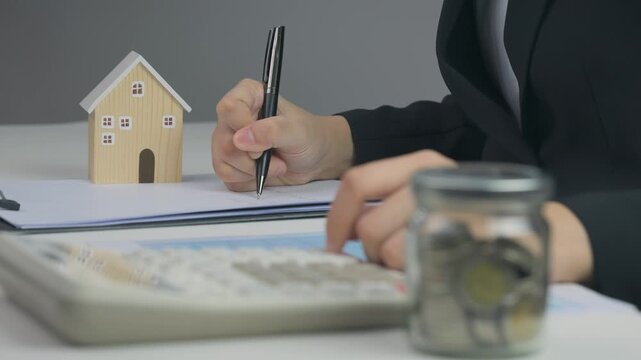 Focused person plans mortgage budget, calculates saving, signs paper, achieving lifelong home ownership goal near wooden house model