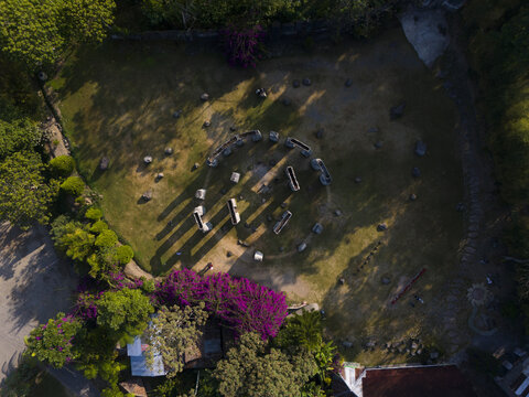 Aerial view of the circular stone arrangement at Warung Kopi Merapi surrounded by lush grass, purple bougainvillea, and trees in Kecamatan Cangkringan, Daerah Istimewa Yogya, Indonesia.