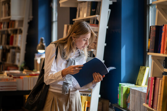 Interested woman booklover reading book album in cozy bookshop, focused female examinees publication, flipping looking through pages, standing near window with stack of books on display. 