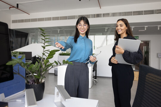 Smiling young 25s Chinese woman office manager welcoming newcomer female employee on first day, giving tour of corporate workplace, pointing at her modern working place, looks friendly and positive