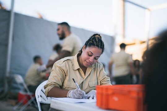 Focused young woman in a beige field uniform writing on a form in a dusty humanitarian camp for aid relief and logistics communication