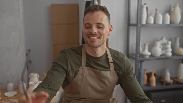 Man in apron holds eyeglasses near his face while smiling with closed eyes in a pottery studio; craft skill contentment.