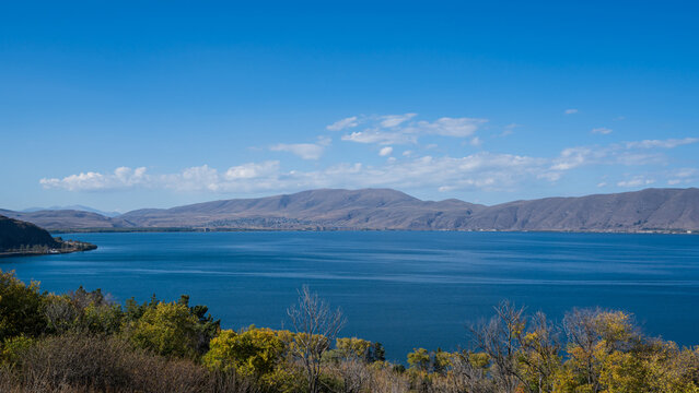 Wide view of Lake Sevan with autumn foliage in the foreground and mountains under a blue sky with soft clouds, Armenia.