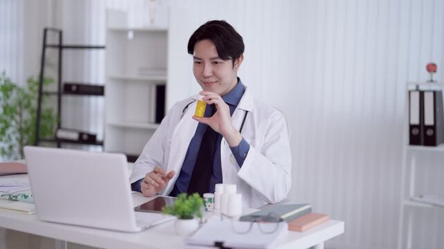 Young Asian doctor presenting medication bottle to laptop camera during telehealth consultation in modern medical office
