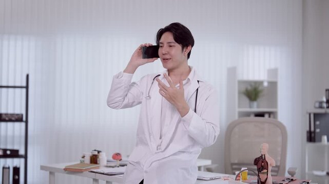 Young Asian doctor speaking on phone during telemedicine consultation in bright modern medical office with copy space