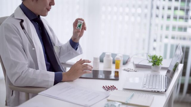 Asian male physician examining medication bottle at modern clinic desk with laptop and medical supplies
