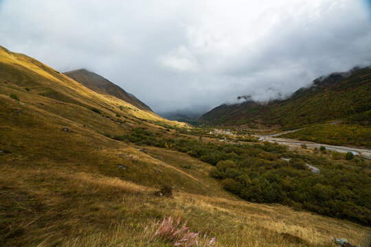 Autumn valley with golden grass slopes and a river winding toward misty peaks near Shkhara glacier above Ushguli, Svaneti, Georgia.