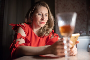 Thoughtful caucasian woman in red dress sitting at a bar or cafe at night, holding a smartphone, with a blurred glass of wine or cocktail in the foreground with dim lighting. © shymar27