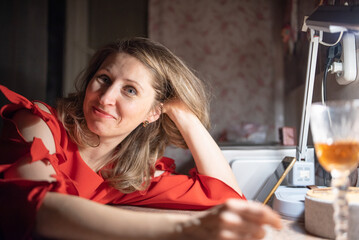 Thoughtful caucasian woman in red dress sitting at a bar or cafe at night, holding a smartphone, with a blurred glass of wine or cocktail in the foreground with dim lighting. © shymar27