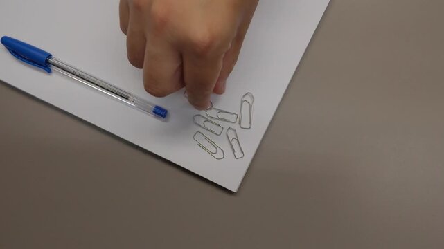 Top view of a hand removing paper clips one by one from a stack of blank papers with a blue pen on an office desk, representing organization and document management.
