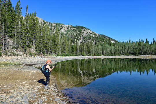 Woman explores shore of second Thomas Lake in Maroon Bells-Snowmass Wilderness in White River National Forest on calm clear summer morning.
