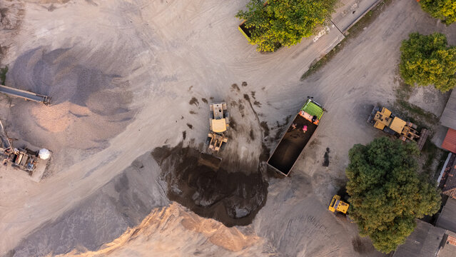 Aerial view of a sand mining site with a yellow front-end loader loading gravel into a truck, surrounded by piles of sand and trees in Klaten, Central Java, Indonesia.