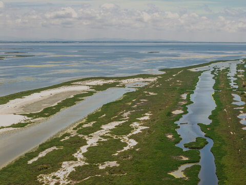 Aerial view of Karavasta Lagoon with winding channels, lush green marshland, sandy banks, and calm blue water under a cloudy sky, Divjake, Fier County, Albania.