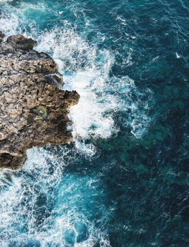 Aerial view of the rugged rocky coastline with crashing white waves and deep blue Mediterranean sea in Binibequer Nou, Illes Balears, Spain.