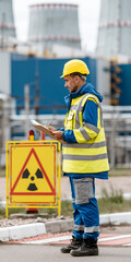 Obraz premium A male worker wearing a yellow hard hat and safety vest stands near a nuclear power plant, holding a tablet and inspecting a radiation hazard sign