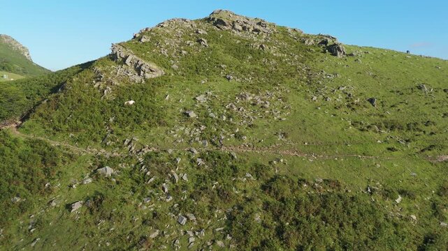 Aerial tracking shot panning right along Zizkuitza mountain in Nouvelle-Aquitaine, France. A lone hiker on a remote, green trail under a clear blue sky, showcasing the scenic, rugged landscape.
