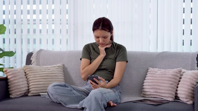 Young Asian woman showing worried expression while reading bad news on smartphone sitting on living room sofa