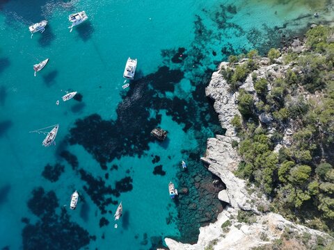 Aerial view of Binibequer Vell with boats anchored in the turquoise waters of the Mediterranean Sea, rocky coastline, and pine trees in Binibequer Nou, Illes Balears, Spain.