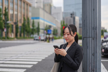 Fototapeta premium 丸の内の歩道でスマートフォンを見る若いビジネスウーマン