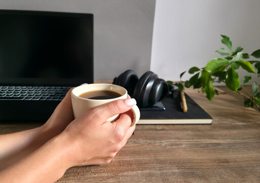 Hands holding a cup of coffee with a laptop, headphones and notebook in the background. Concept of a work break and end of workday