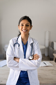 Smiling South Asian female doctor in medical coat standing with arms crossed, posing for camera in clinic examination room, represents professionalism, healthcare services, patient care, portrait