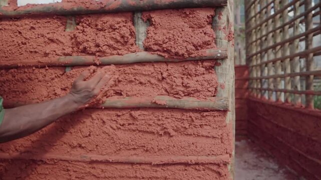 Close up of a construction worker's hands applying a mixture of mud and straw to a eucalyptus wood frame, building a natural house