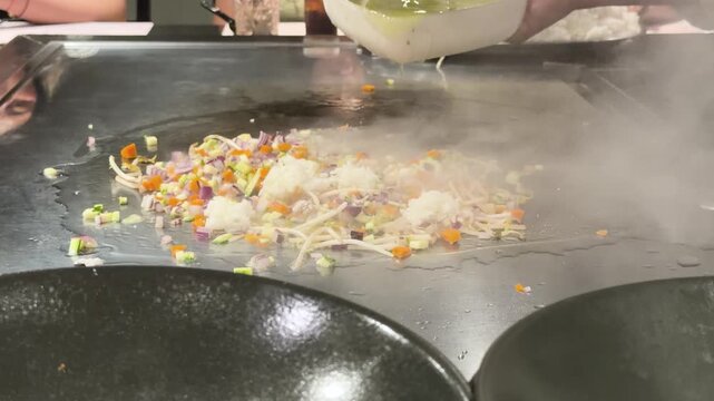 Chef prepares oriental style fried rice with vegetables on hot flat top griddle, drizzling oil over food