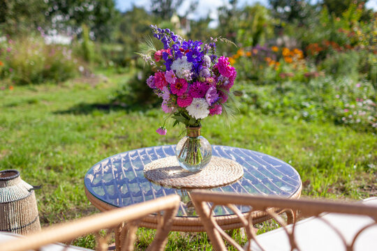 Close up of flower arrangement put on glass table in summer garden. Lounge zone with furniture and bouquet of fresh blooms in vase