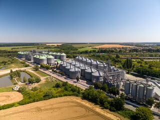 High angle view captures massive agricultural storage plant with rows of steel silos next to rural green fields under bright sun. © Pasha