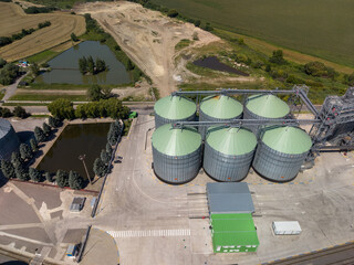 Straight down viewpoint of six circular metal grain silos with light green tops built next to artificial ponds on sunny day. © Pasha