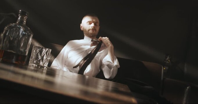 Tired man loosens his tie while sitting at a table with whiskey and eventually takes a drink.