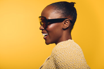 Smiling African woman with dark skin wearing large sunglasses and a knitted sweater against yellow background. Profile portrait captures joy, fashion, and confident mood with bright studio lighting. © SHOTPRIME STUDIO