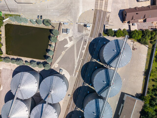 Straight down viewpoint of metal grain silos arranged near railway tracks and artificial rectangular water pond on sunny day. © Pasha