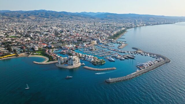 Aerial view of Limassol Marina in Cyprus. Curved harbour walls, calm blue water, yachts and the coastal city skyline create a scenic Mediterranean waterfront panorama.