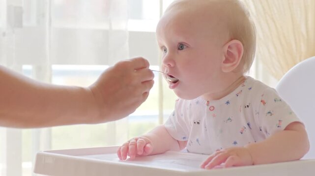 Slow motion: feeding a baby from a spoon. Mom feeds a 9 month baby with fruit puree from a spoon, close-up, high key. Meal time. Little boy eats sitting on baby's chair. 