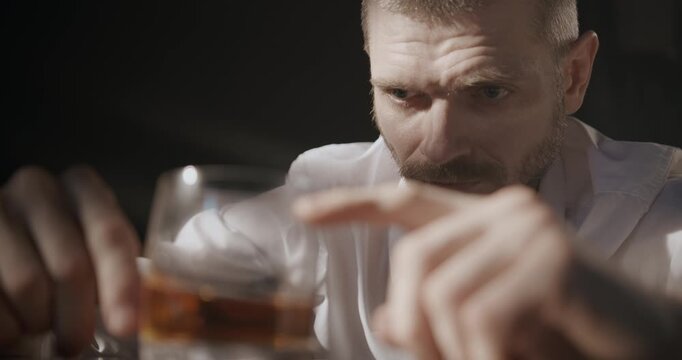  An anxious man nervously moves a glass of whiskey back and forth on the table while staring at it with a tense expression.