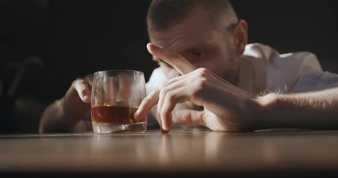 A hesitant man moves a glass of whiskey back and forth on the table while staring at it with a troubled expression.