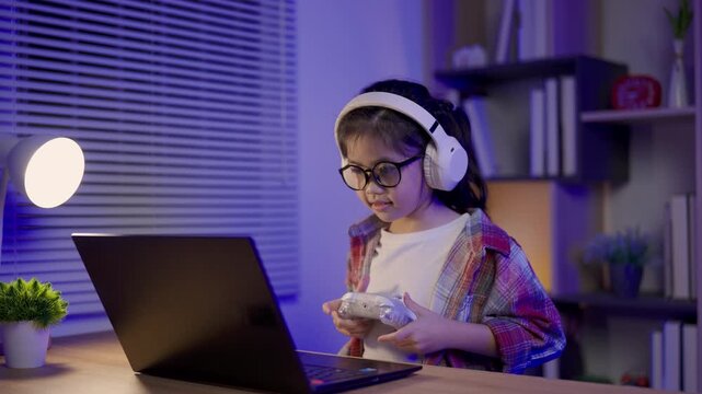 Young child wearing glasses and headphones holding game controller concentrating while playing video game at home living room in evening