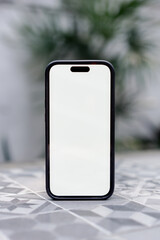 Modern smartphone stands upright on a tiled surface. Its screen is blank, ready for your content. Soft-focus plants create a calm, natural backdrop. mockup phone   © faizal