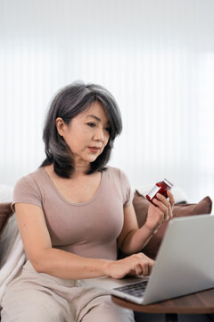 Woman holding pill bottle and using laptop computer searching for information about  prescription medicine while sitting in livingroom at home .