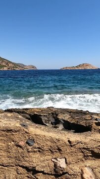 Camera movement reveals  a small rocky islet in the Greek sea. Vertical slow motion video. Clear blue sky at the background.