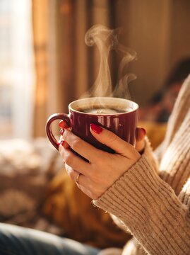 Closeup of a womans hands holding a steaming mug of hot coffee, wearing a cozy beige knitted sweater in a warm, sunlit indoor setting